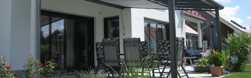 Terrassendächer Alu Bremen: Eine Überdachung mit Unterglasmarkise in Anthrazit mit vor Sonnenlicht geschütztn Gartenmöbel darunter.