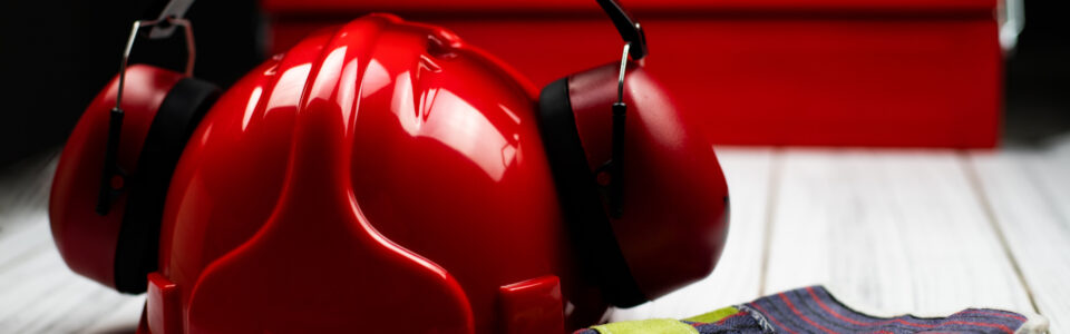 Gehörschutz Bremen: Ein roter Helm, auf dem ein ebenfalls roter Gehörschutz montiert ist, daneben Arbeitshandschuhe